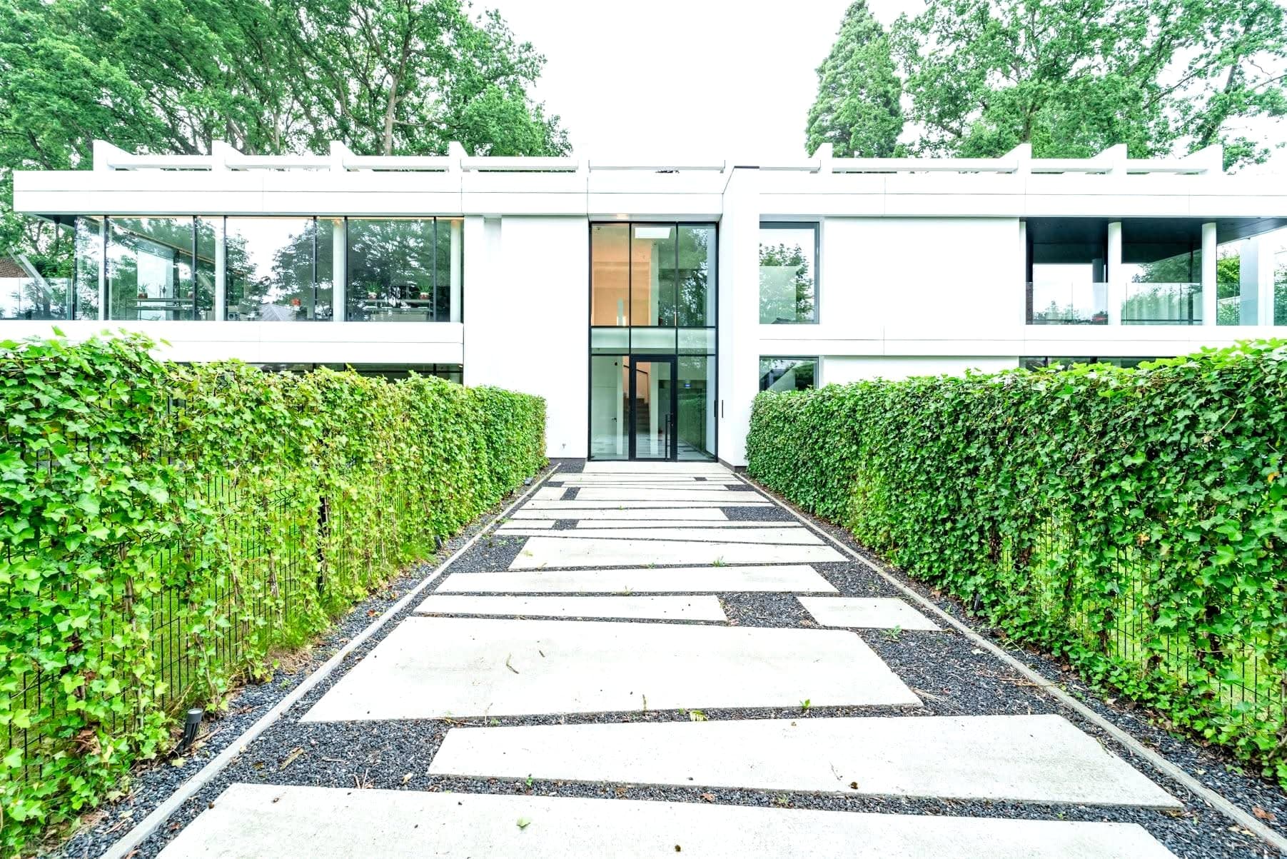Allée d'accès de la villa : dalles de béton sur gravier noir entre deux haies, entrée vitrée double hauteur.